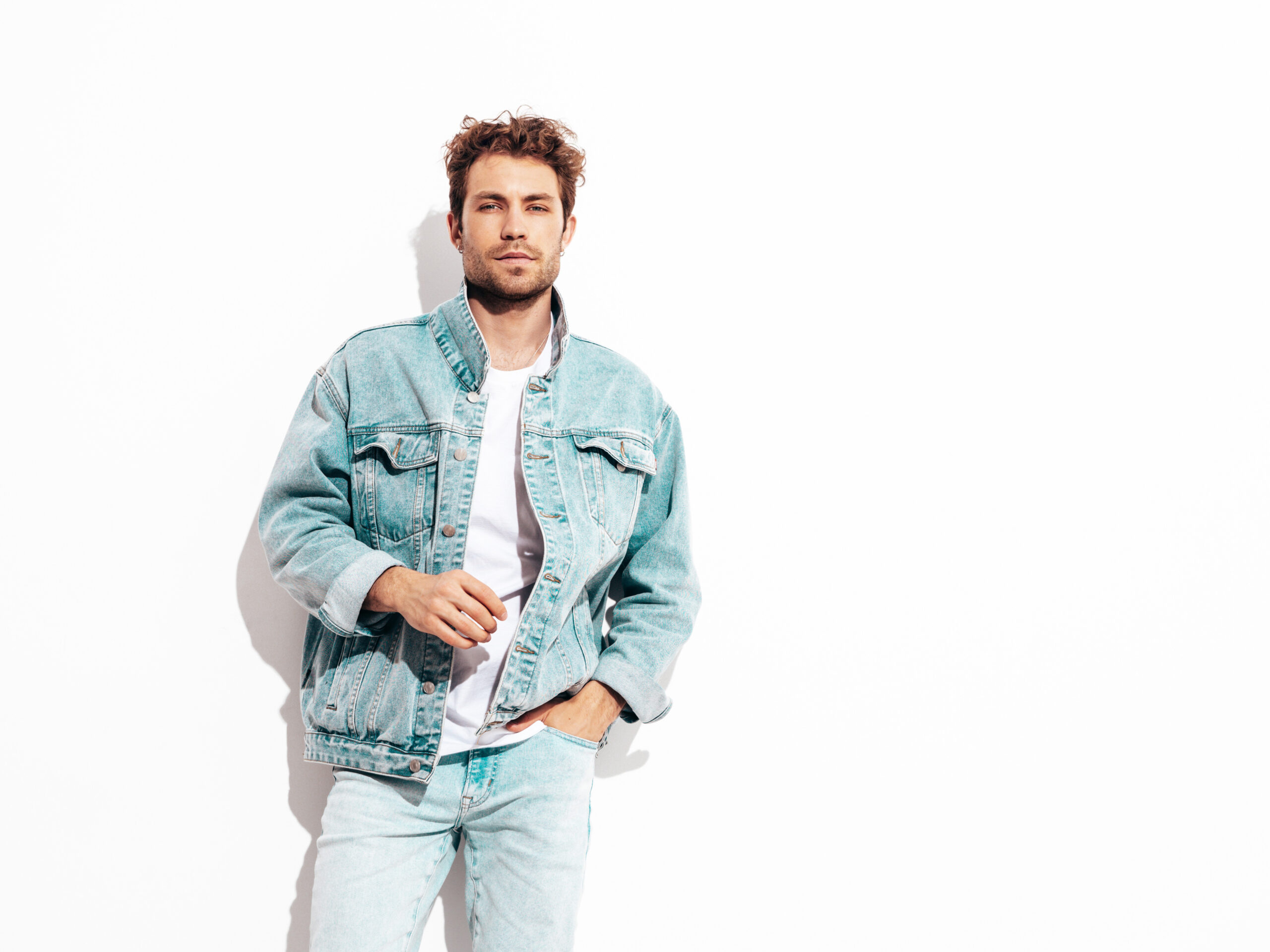 Portrait of handsome confident stylish hipster lambersexual model with curly hairstyle. Sexy man dressed in jeans jacket. Fashion male isolated on white wall in studio